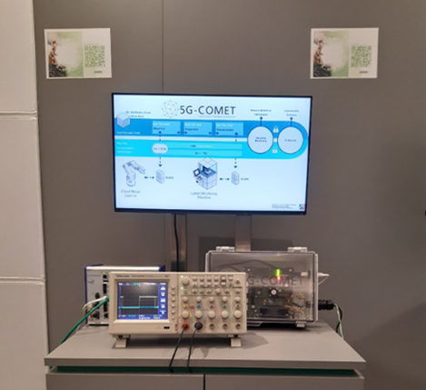 A display showcasing a 5G technology project titled "5G-COMET." In the center, a large screen presents a diagram with blue and grey icons illustrating various components such as Cloud Radio Gateways and Laser-Interfacing Modules. Below the screen, an arrangement of electronic equipment is visible, including an oscilloscope on the left and a more complex instrumentation device in the center, enclosed in a transparent casing. The background is a plain grey wall with two QR code posters affixed to the top corners. The overall setup suggests a focus on advanced technology and research in telecommunications.
